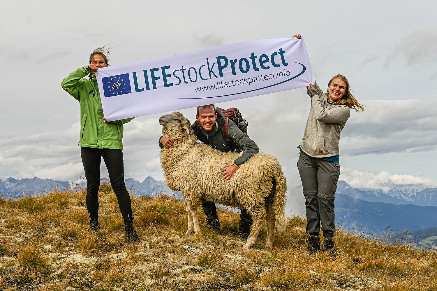 Schaf, von einer Person umarmt sowie zwei stehende Personen, die über dem Schaf eine Fahne mit der Aufschrift LIFEstockProtect halten.